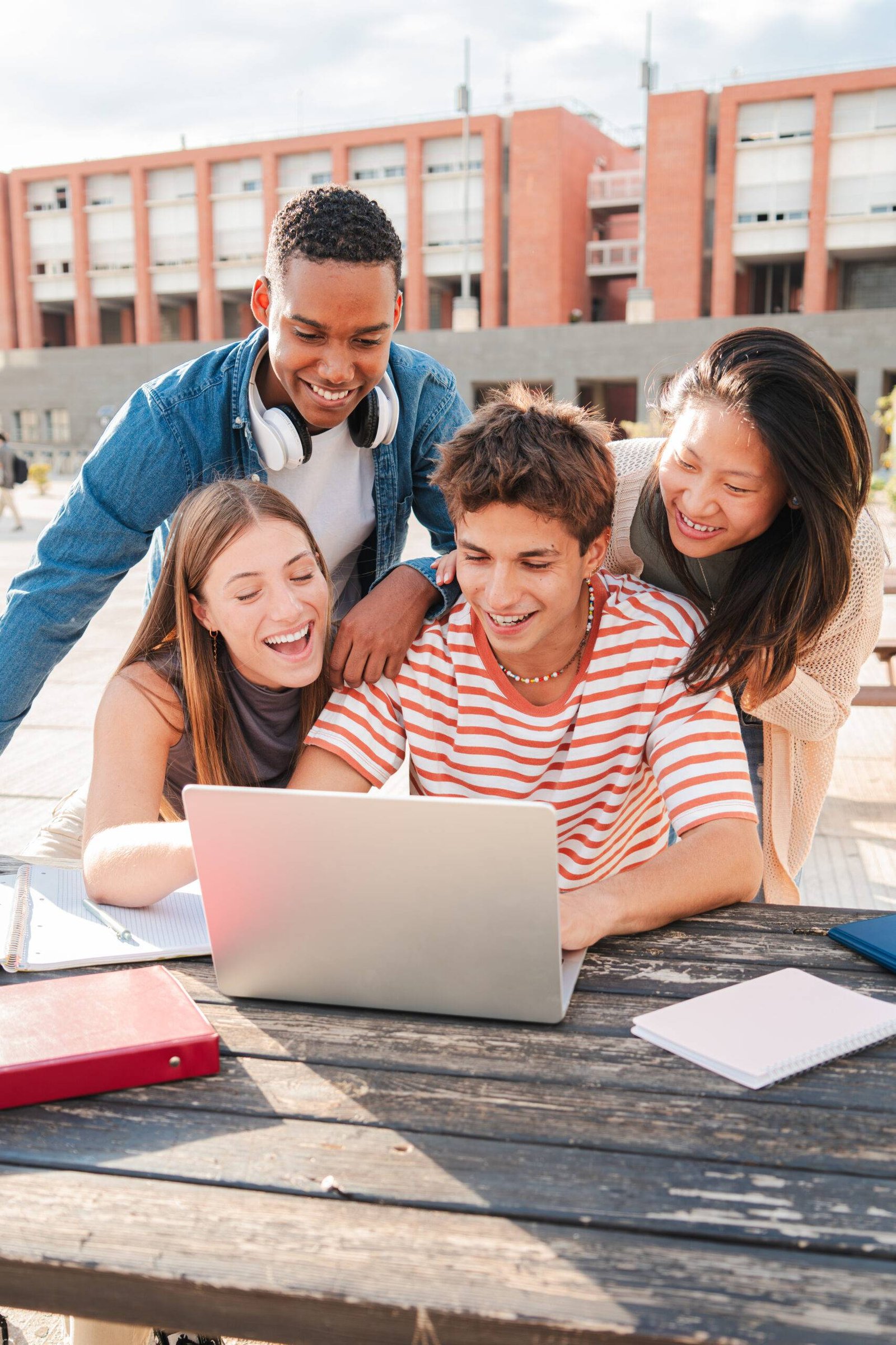 Vertical. Group of young classmates studying together and preparing a exam using a laptop, browsing on internet at university campus. Teenage students smiling and doing the homeworks with a computer